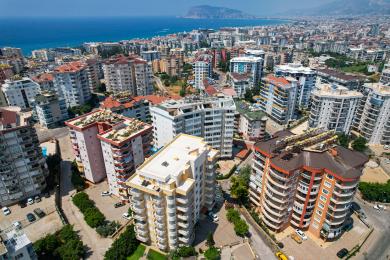 Sala de estar luminosa do apartamento Alanya com cozinha aberta, pisos de cerâmica, cadeiras de vime e sofá cinza confortável.