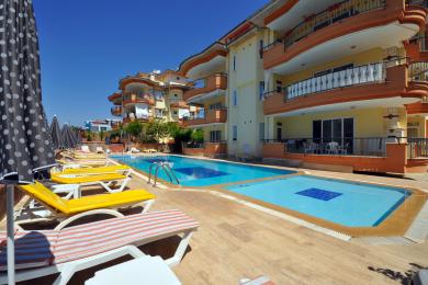Alanya balkon met wicker meubilair kijkt uit over een helderblauw zwembad en zonnige buurt.