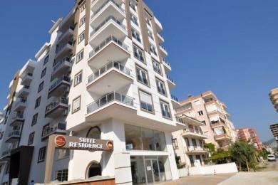 Alanja apartman dnevni boravak sadrži modernu kuhinju, trpezarijski set, teksturirani zid i udoban beli kauč.