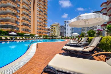 Yorug' Alanya xonadonining yashash maydonida zamonaviy oq ovqatlanish stoli, zamonaviy oshxona va qulay kulrang divan mavjud.
