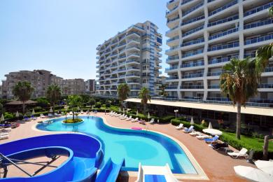 Vista de la propiedad de Alanya: piscina estilo resort con toboganes, rodeada de palmeras y edificios de apartamentos, ambiente soleado.
