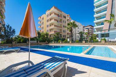 Een balkon uitzicht vanaf een appartement in Alanya toont een met palmen omzoomde straat, gebouwen en verre bergen onder een heldere, zonnige hemel.