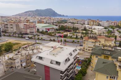 Das Wohnzimmer der Alanya Immobilie verfügt über eine moderne Küche, ein graues Sofa und helles, natürliches Licht durch große Fenster.