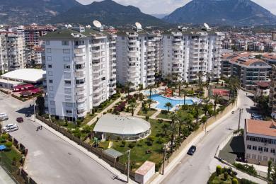 Alanya'da satılık mülk: Bej renkli koltuğa, fayans zemine ve modern beyaz mutfağa sahip, aydınlık, açık planlı oturma odası.