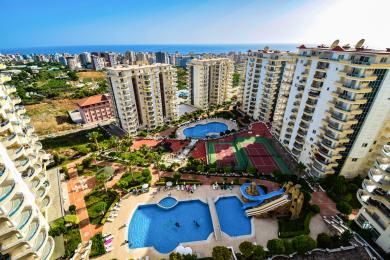 Alanya Immobilien Balkonblick zeigt Pools, Tennisplätze und üppige Landschaft unter hellem Sonnenlicht und schafft eine Resort-Atmosphäre.