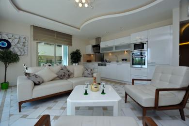 El luminoso salón y la cocina del apartamento en Alanya cuentan con muebles blancos, suelos de mármol y electrodomésticos modernos.