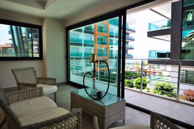 Lys Alanya balkon med kurvemøbler, glasbord og skydedøre med udsigt over moderne bygninger.