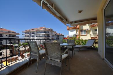 Alānijas īpašuma balkons ar pītiem mēbeļiem, flīžu grīdu un spilgtu dienasgaismu, kas paveras uz tuvējām ēkām.