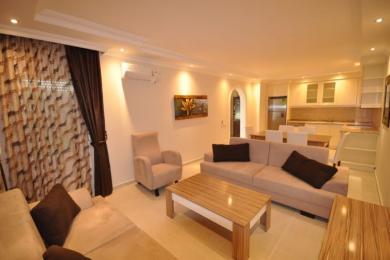 Salon de propriété à Alanya avec des canapés neutres, un revêtement de sol en carrelage et une cuisine américaine avec un éclairage chaleureux et accueillant.