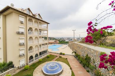 Alanya fastighetsvy som visar en pool, fontän och balkonger med en stads- och havsbakgrund.