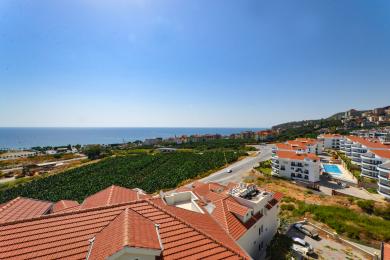 Alanya əmlakının qırmızı kirəmitli damları, bol yaşıllıqları və aydın mavi səma altında parıldayan Aralıq dənizini nümayiş etdirən mənzərəsi.