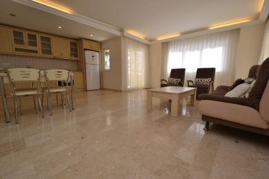 The bright, open-plan living area of this Alanya property features a kitchen, marble floors, and comfortable seating near sheer-curtained windows.