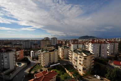 Alanya mulkining bulutli osmon ostida shahar silueti, ko'p qavatli uylar va O'rta er dengizini namoyish etuvchi ko'rinishi.