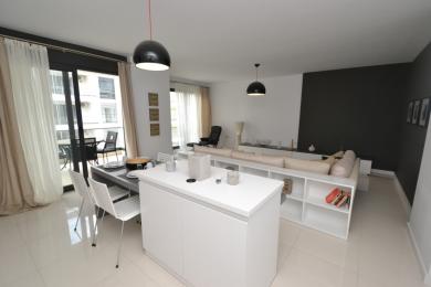 Svetlá otvorená obývacia izba apartmánu v Alanyi ponúka moderný kuchynský ostrov, prístup na balkón a elegantný minimalistický dizajn.