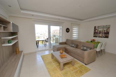 Šviesi svetainė Alanijoje su smėlio spalvos sofa, valgomojo zona, išėjimu į balkoną ir neutraliomis plytelėmis.