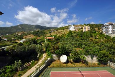 Vista da propriedade em Alanya, mostrando um campo de ténis com vista para a montanha e edifícios sob um céu azul.
