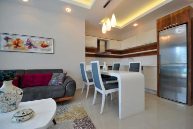 Yorug' Alanya xonadonining yashash maydonida zamonaviy oq ovqatlanish stoli, zamonaviy oshxona va qulay kulrang divan mavjud.