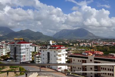 Alanya Immobilien Balkonansicht mit Stadtgebäuden und fernen Bergen unter einem teilweise bewölkten Himmel.