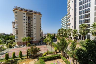 Vue d'une propriété à Alanya présentant des immeubles d'appartements modernes, une végétation luxuriante, un court de tennis et une aire de jeux sous un ciel bleu clair.