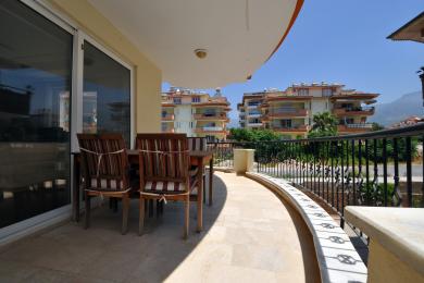 Balkón nehnuteľnosti Alanya s drevenou jedálenskou súpravou, dlaždenou podlahou a slnečnou, uvoľnenou atmosférou.