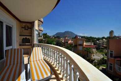 Alanya-Immobilienbalkon mit gestreiften Liegen, Steinkamin und atemberaubendem Bergblick unter einem klaren blauen Himmel.