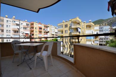 Güneşli Alanya balkonu, fayans zemin, beyaz bir masa seti ve yakındaki apartman binalarının manzarasına sahip.