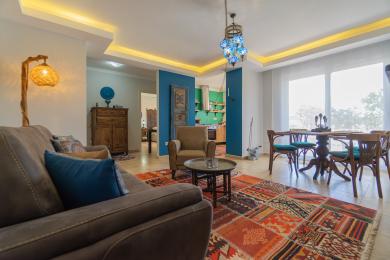 Alanya eign stofa með litríku teppi, bláum áherslum, hlýrri lýsingu og opnu eldhúsi sem skapar notalega stemningu.