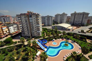 Alanya mulkidan ko'rinish: Suralar bilan jihozlangan kurort uslubidagi hovuz, atrofida palma daraxtlari va ko'p qavatli uylar, quyoshli muhit.