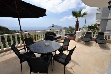 Balkón nehnuteľnosti Alanya s čiernym prúteným nábytkom, vírivkou a panoramatickým výhľadom na more za jasného denného svetla.