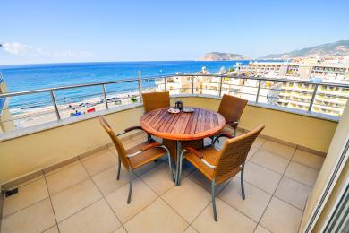 Balcone di una proprietà ad Alanya con un tavolo di legno, sedie in vimini e splendida vista sul mare sotto la luce del sole.