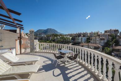 Alanya mulkidagi kafel qoplamali balkon, nafis panjara, dam olish stullari va yorqin osmon ostidagi ajoyib tog 'manzarasi.
