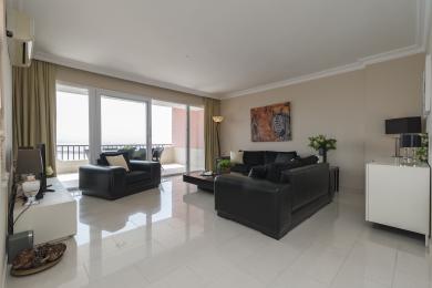 Soggiorno di un appartamento ad Alanya con divani in pelle nera, pavimenti in piastrelle lucide e accesso al balcone, creando un'atmosfera luminosa e ariosa.