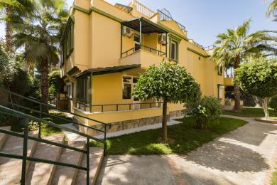 Alanya daşınmaz əmlakının eksteryeri: Eyvanları, yaşıl haşiyələri, bol bağı və günəşli Aralıq dənizi atmosferi olan sarı villa.
