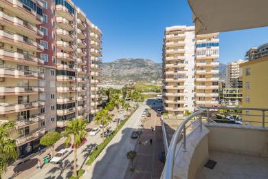 Een balkon uitzicht vanaf een appartement in Alanya toont een met palmen omzoomde straat, gebouwen en verre bergen onder een heldere, zonnige hemel.