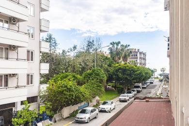 Alanya mülkündən balkon mənzərəsi, günəşli bir gündə sıx yaşıl ağaclara və park edilmiş avtomobillərə baxır.