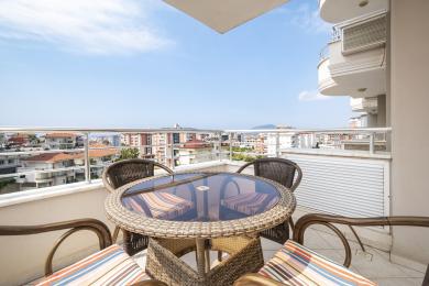 Alanya'da hasır mobilyalar, cam masa ve parlak, güneşli bir gökyüzü altında panoramik şehir manzarası sunan balkonlu bir mülk.