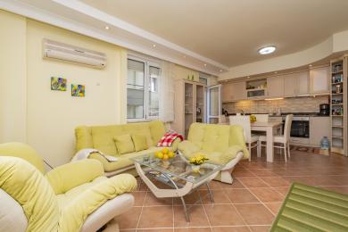 Alanya Immobilie Wohnzimmer mit gelben Sofas, Fliesenböden und einer offenen Küche, die eine helle und einladende Atmosphäre schafft.