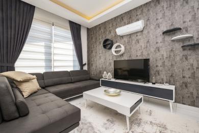 Alanya fastighet vardagsrum har en grå soffa, texturerad tapet och moderna vita möbler under varm belysning.