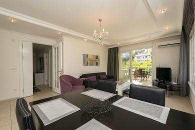 Alanya Immobilie Wohnzimmer mit Kronleuchter, dunklem Holzesstisch und Zugang zum Balkon, wodurch eine helle, luftige Atmosphäre entsteht.