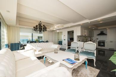 Sala de estar luminosa em Alanya com sofás brancos, lustre preto e cozinha de plano aberto com pisos de mármore e vista para o mar.