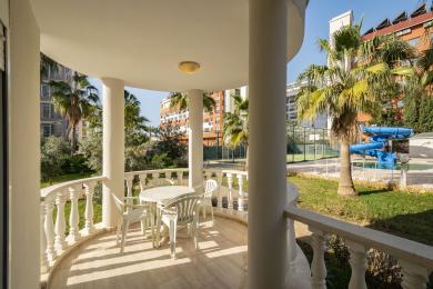 Alanya-Immobilienbalkon mit weißen Säulen, Fliesenboden und Sitzgelegenheiten im Freien mit Blick auf üppige tropische Anlagen.