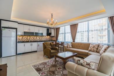 Jasny apartament w Alanyi, salon z nowoczesną białą kuchnią, kafelkową podłogą i wygodnymi siedziskami w neutralnych odcieniach, oświetlony delikatnym światłem.