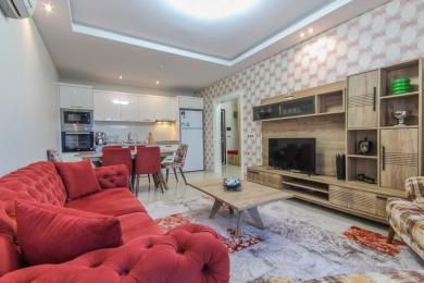 Salon de propriété à Alanya avec un canapé rouge, une cuisine moderne et un meuble TV en bois clair, créant une atmosphère chaleureuse.