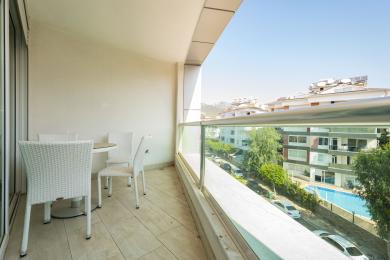Bright Alanya balcony featuring white wicker furniture, tile flooring, and a glass railing with city views.