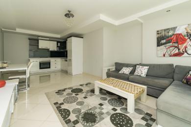 De heldere woonkamer van dit appartement in Alanya beschikt over een moderne keuken, een grijze bank, een vloerkleed met patroon en glanzende tegelvloeren.