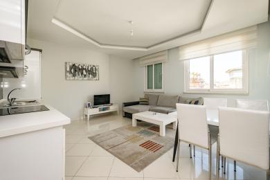 Světlý obývací prostor apartmánu v Alanyi nabízí šedou rohovou sedačku, bílý jídelní set a dlažbu.