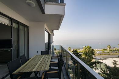 Balcone di proprietà ad Alanya con tavolo in legno, sedie, ringhiera in vetro e splendida vista sul mare sotto la luce del sole.