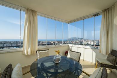 Balcón luminoso en Alanya con paredes de cristal, vistas a la ciudad, muebles de mimbre y cortinas neutras que crean un ambiente relajante.