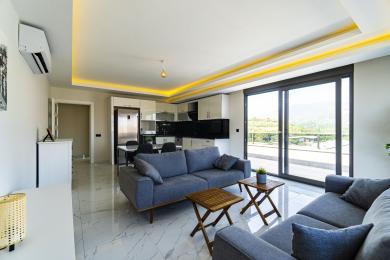 Alanya mulkidagi yashash xonasida zamonaviy oshxona, marmar pollar, kulrang divanlar va balkon eshiklaridan kirib kelayotgan yorqin tabiiy yorug'lik mavjud.