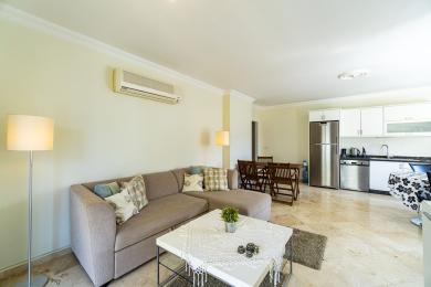 Alanijos nuosavybės svetainė su marmurinėmis grindimis, pilka sofa ir šviesia, modernia virtuvės zona.